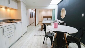 A modern hotel room with a kitchenette, a dining area with a round table and chairs, a bed with a headboard, and a large mirror on the wall.