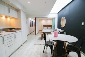 A modern hotel room with a kitchenette, a dining area with a round table and chairs, a bed with a headboard, and a large mirror on the wall.