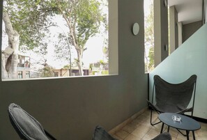 A room with a view of trees and buildings, a chair, a small table, and a staircase.