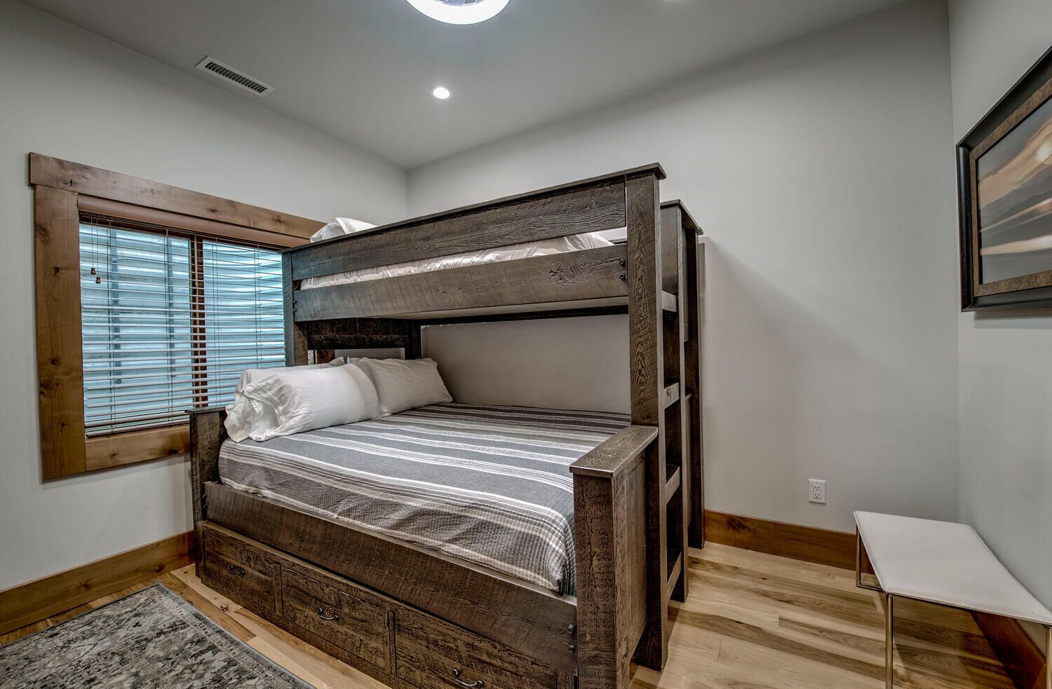 Un lit superposé avec une structure en bois, un matelas à rayures grises et des oreillers blancs. Il y a une petite table blanche à côté du lit, une fenêtre avec des stores et un plancher en bois.