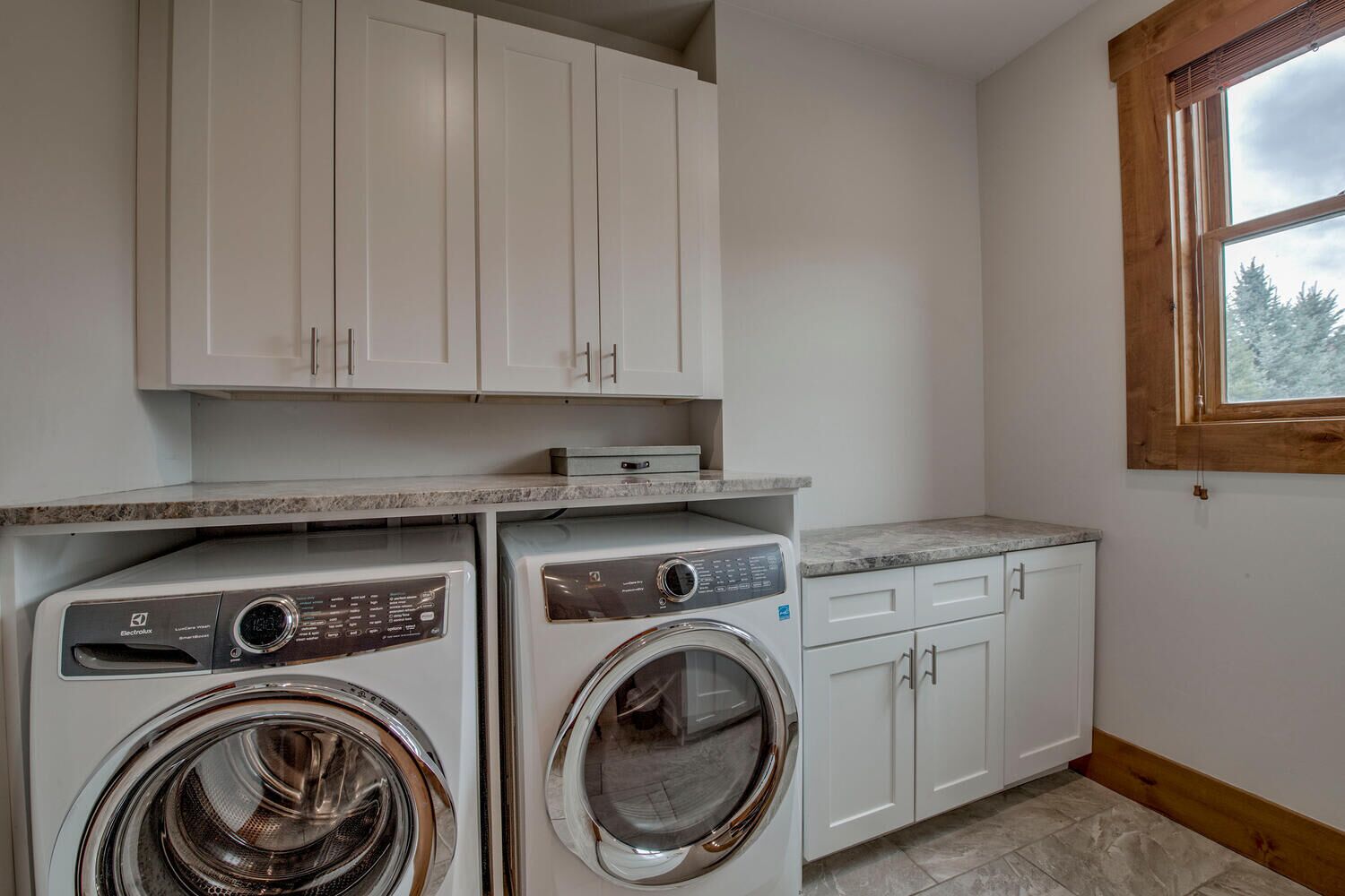 A laundry room with two front-loading washing machines, a countertop, cabinets, and a window with a view of trees.