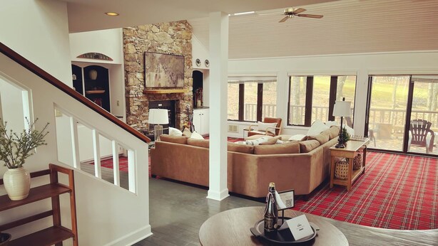 Uma sala ampla com lareira de pedra, uma janela grande com vista para um deck e uma mesa de centro redonda com uma garrafa e apoio para copos.