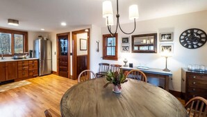 A dining area with a round wooden table, chairs, a chandelier, and a kitchen in the background. - Centrally Located Historical Home (Lee)