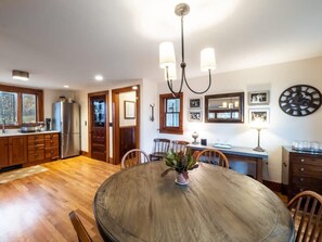A dining area with a round wooden table, chairs, a chandelier, and a kitchen in the background. - Centrally Located Historical Home (Lee)