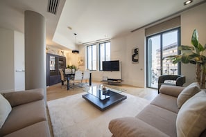 Uma sala de estar moderna com mesa de centro de vidro, sofás bege, área de jantar com um aparador escuro e cadeiras, televisão e janelas amplas.