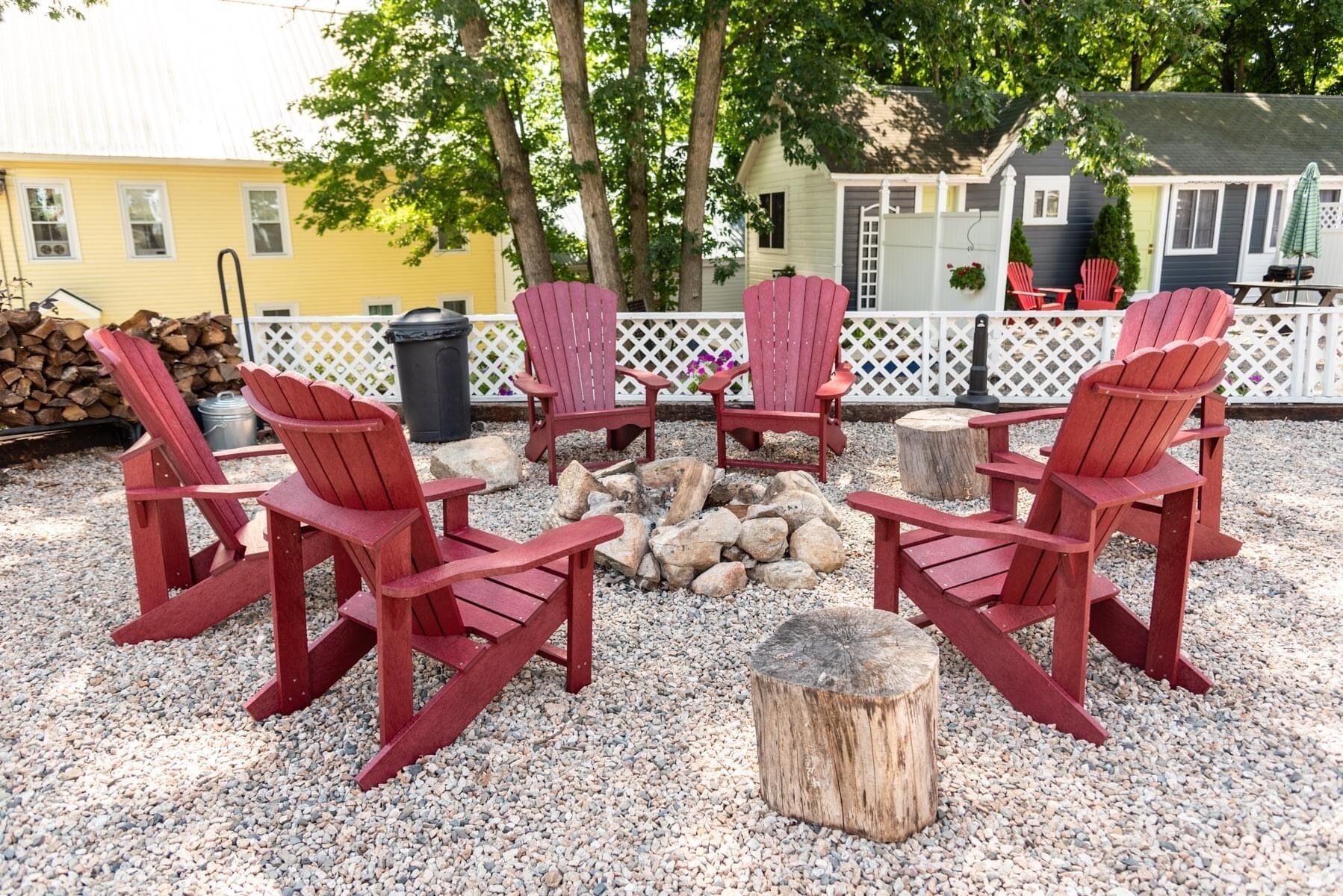 A fire pit surrounded by red Adirondack chairs, with a gravel ground and a white lattice fence in the background.
