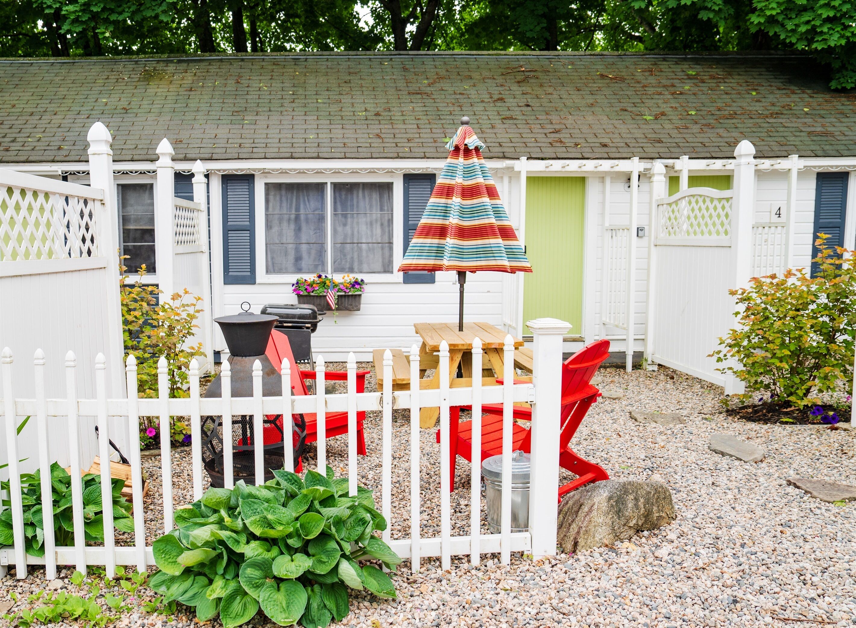 A backyard with a white picket fence, a wooden deck, a colorful striped umbrella, and a grill.