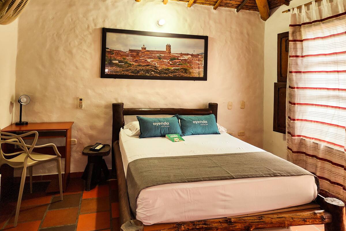 A hotel room with a bed, a desk, a chair, a nightstand, a picture on the wall, and a window with curtains.