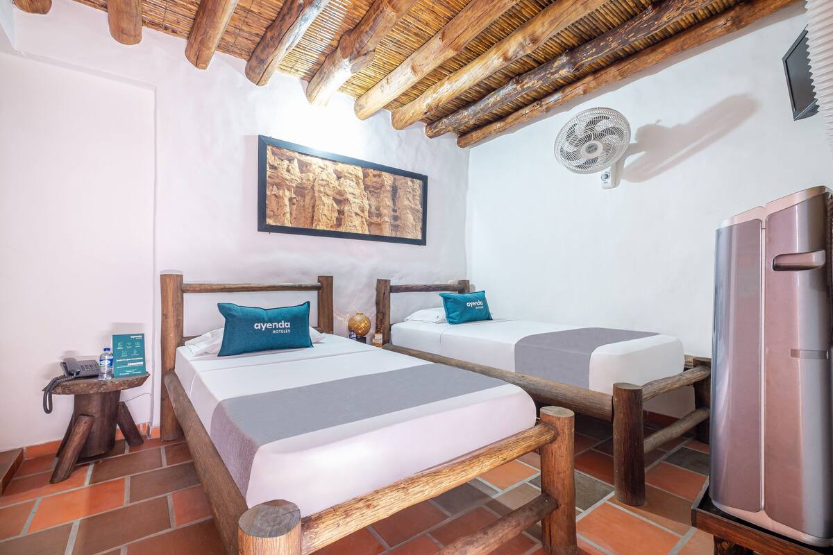 A room with two beds, a wooden ceiling, a wall-mounted fan, and a refrigerator.