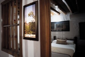 Une photo encadrée représentant un arbre, avec une étiquette indiquant « Los Santos », est accrochée à un mur dans une pièce qui contient un lit et une fenêtre dotée de volets en bois.