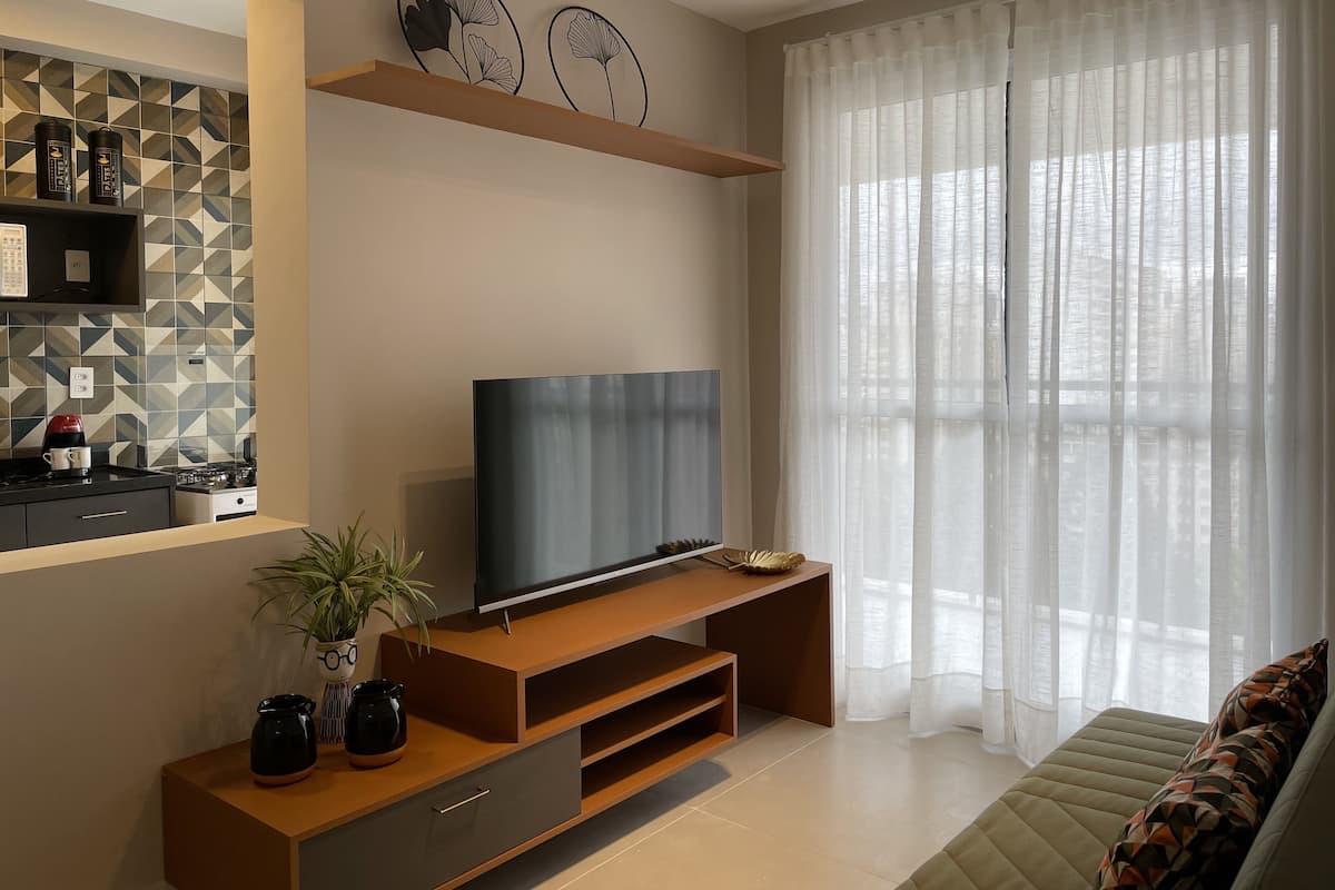 A modern living room with a flat-screen TV on a wooden stand, a kitchen area with a coffee maker, and a large window with sheer curtains.