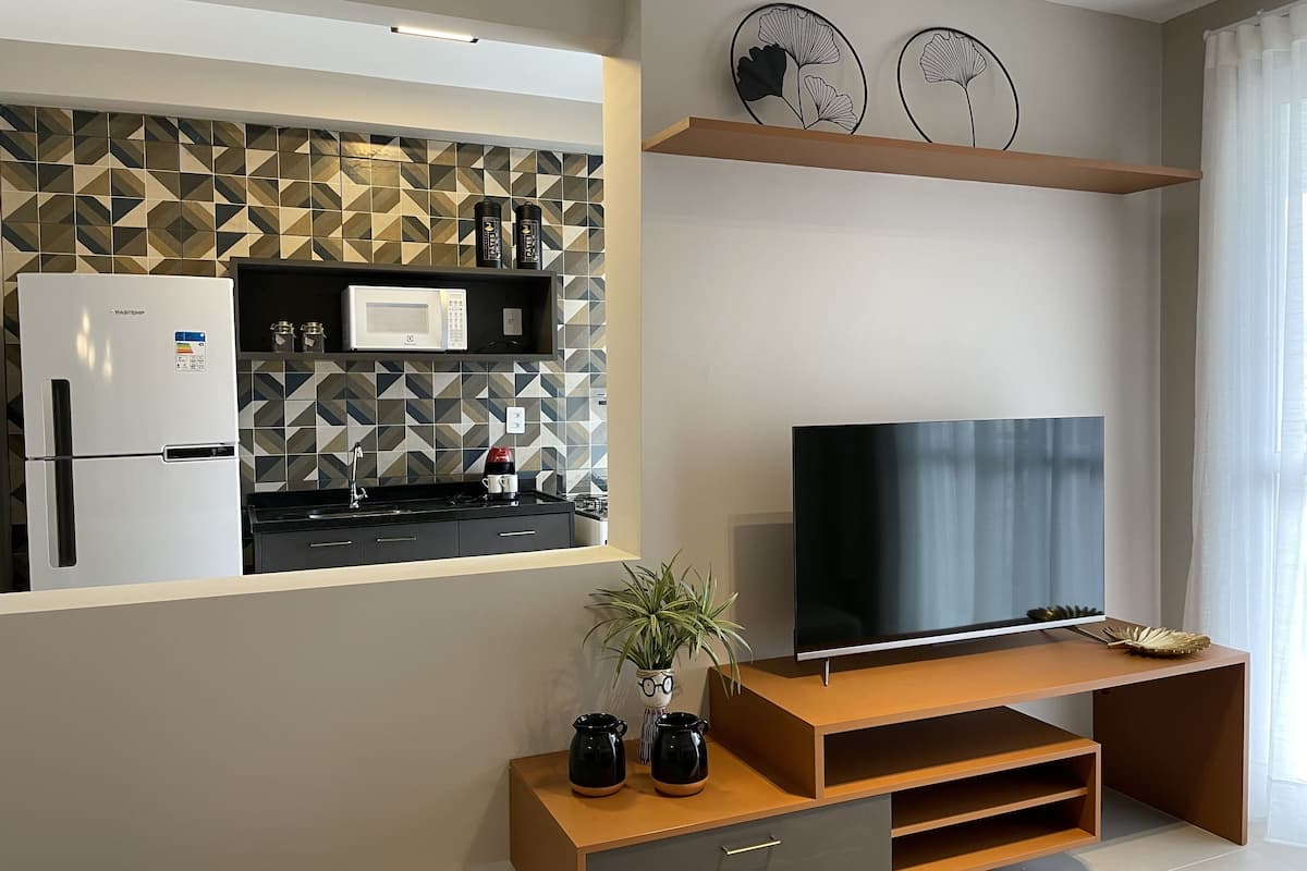 A modern kitchen with a refrigerator, microwave, and coffee maker. A TV is mounted on a wooden stand with a plant beside it.
