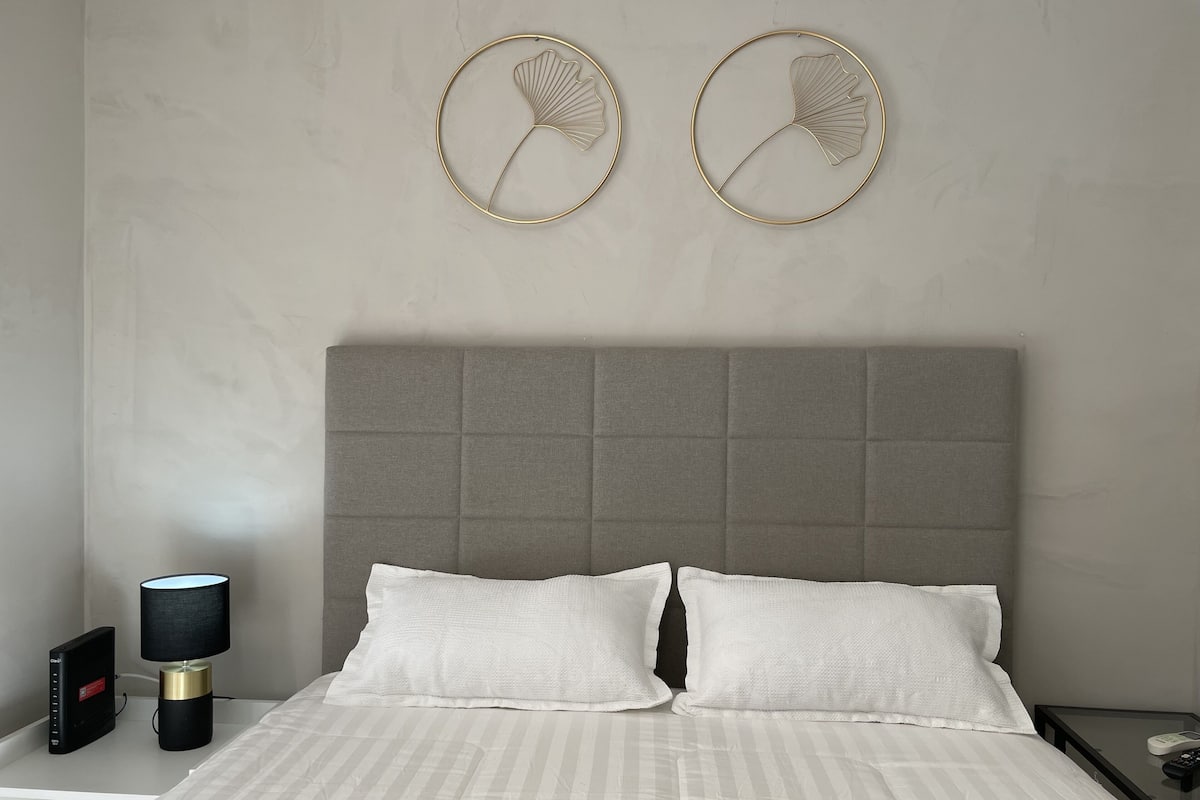 A modern bedroom with a grey upholstered headboard, a white striped bedspread, and two wall-mounted decorative items.