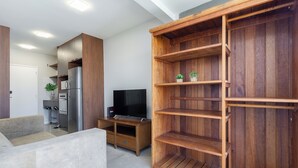 A modern living room with a wooden entertainment unit, a flat-screen TV, and a built-in kitchen area. - Akile Central Park (São Paulo)