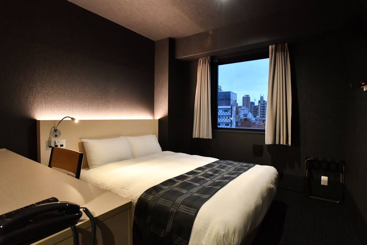 A hotel room with a bed, a desk, a telephone, a window with curtains, and a city view.