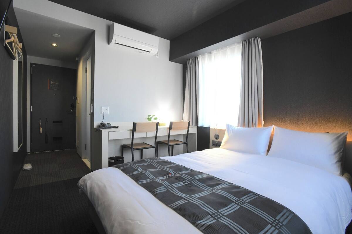 A hotel room with a bed, a desk, chairs, a window with curtains, and an air conditioning unit.