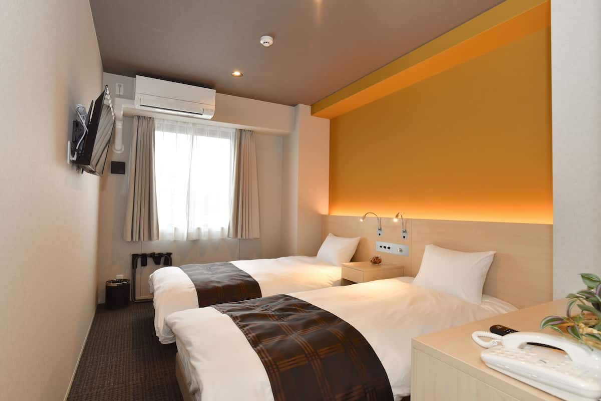 A hotel room with two beds, a bedside table with a phone, a wall-mounted TV, and an air conditioning unit.