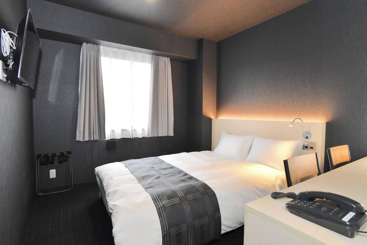 A hotel room with a bed, a desk with a telephone, a lamp, a TV mounted on the wall, and a window with curtains.