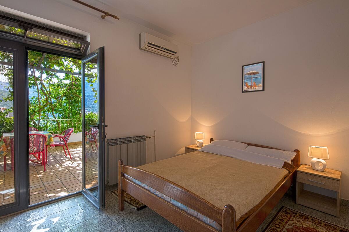 A bedroom with a bed, a nightstand, a wall-mounted air conditioner, a window with a view of greenery, and a small table with chairs outside.