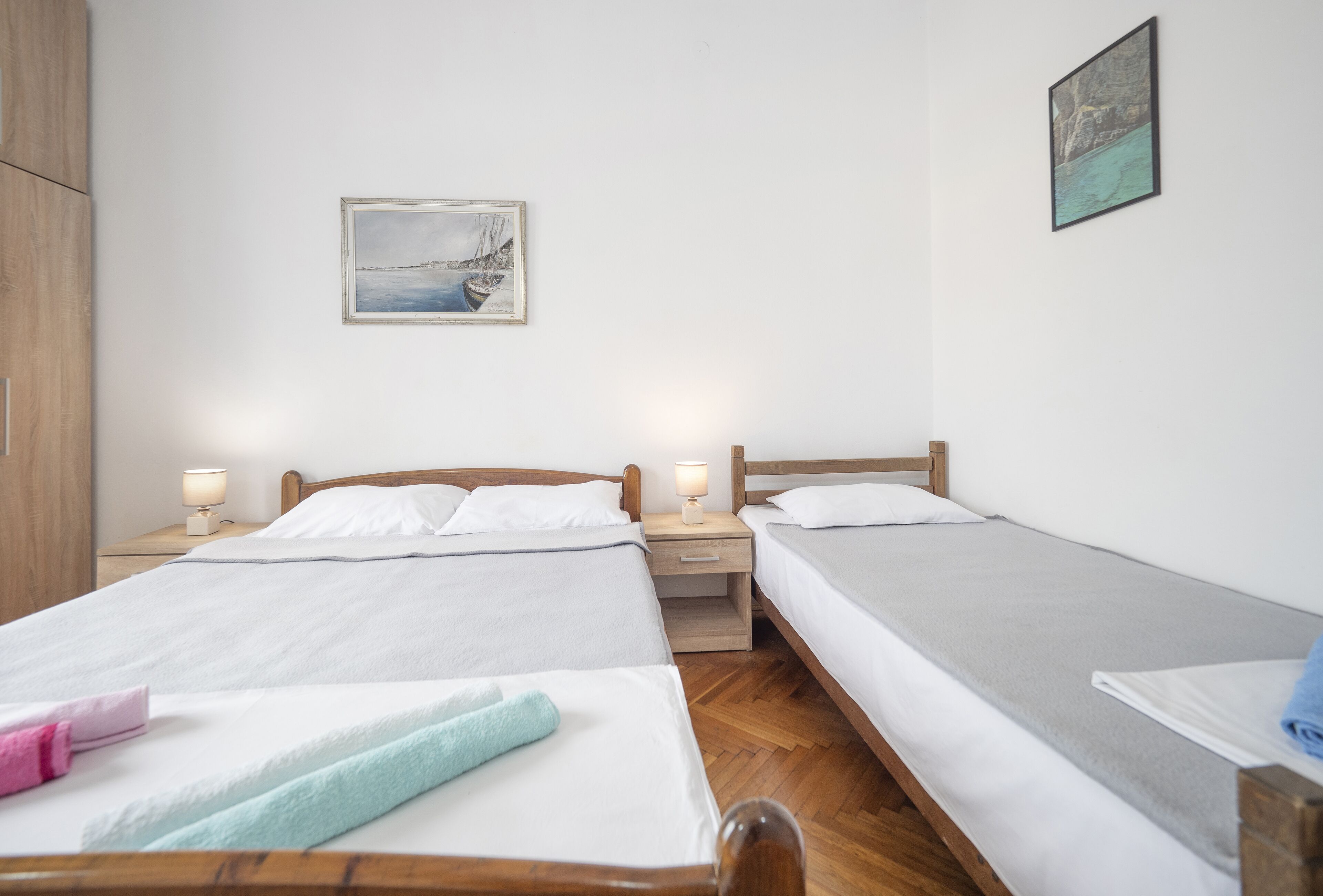 A room with two beds, wooden headboards, white linens, a nightstand with a lamp, and a framed picture on the wall.