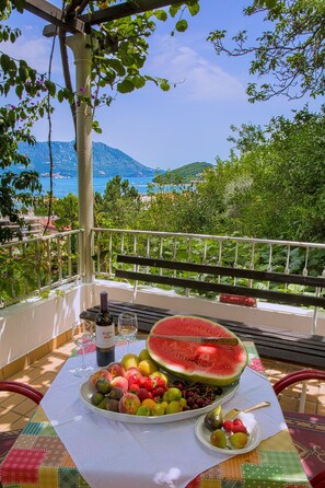 A table with a fruit platter, a bottle of wine, and a watermelon on a balcony with a view of the sea and mountains. - Madra rooms & apartments (Budva)