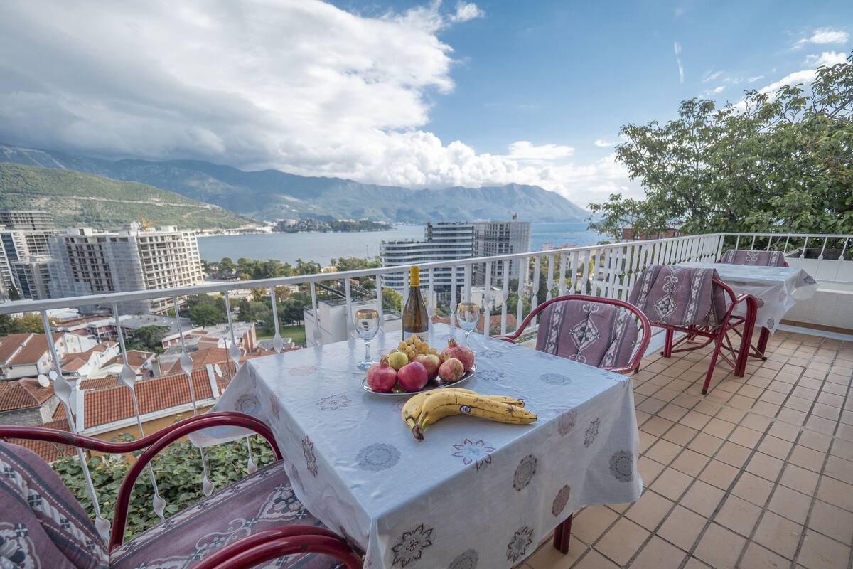 A balcony with a table set for two, featuring a tablecloth with a floral pattern, a bottle of wine, and a plate of fruit.