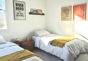 Une chambre avec deux lits simples, une petite table de chevet avec une lampe, une étagère avec des objets décoratifs et des tableaux encadrés au mur.