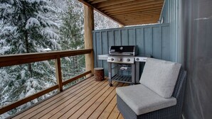Une terrasse couverte avec un barbecue et un fauteuil confortable, d’où l’on peut admirer les arbres couverts de neige.