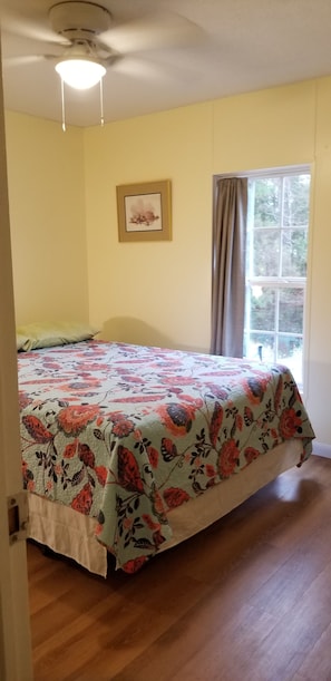A bedroom with a floral bedspread, a ceiling fan, a window with curtains, and a framed picture on the wall.