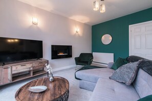 A modern living room with a flat-screen TV, a fireplace, a green armchair, and a grey sofa.