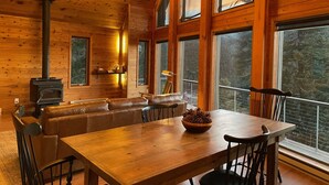 A wooden cabin interior with a large dining table, leather seating, a fireplace, and large windows offering a view of the forest. - Mountain Retreat with Hot Tub Near State Park (Golden)