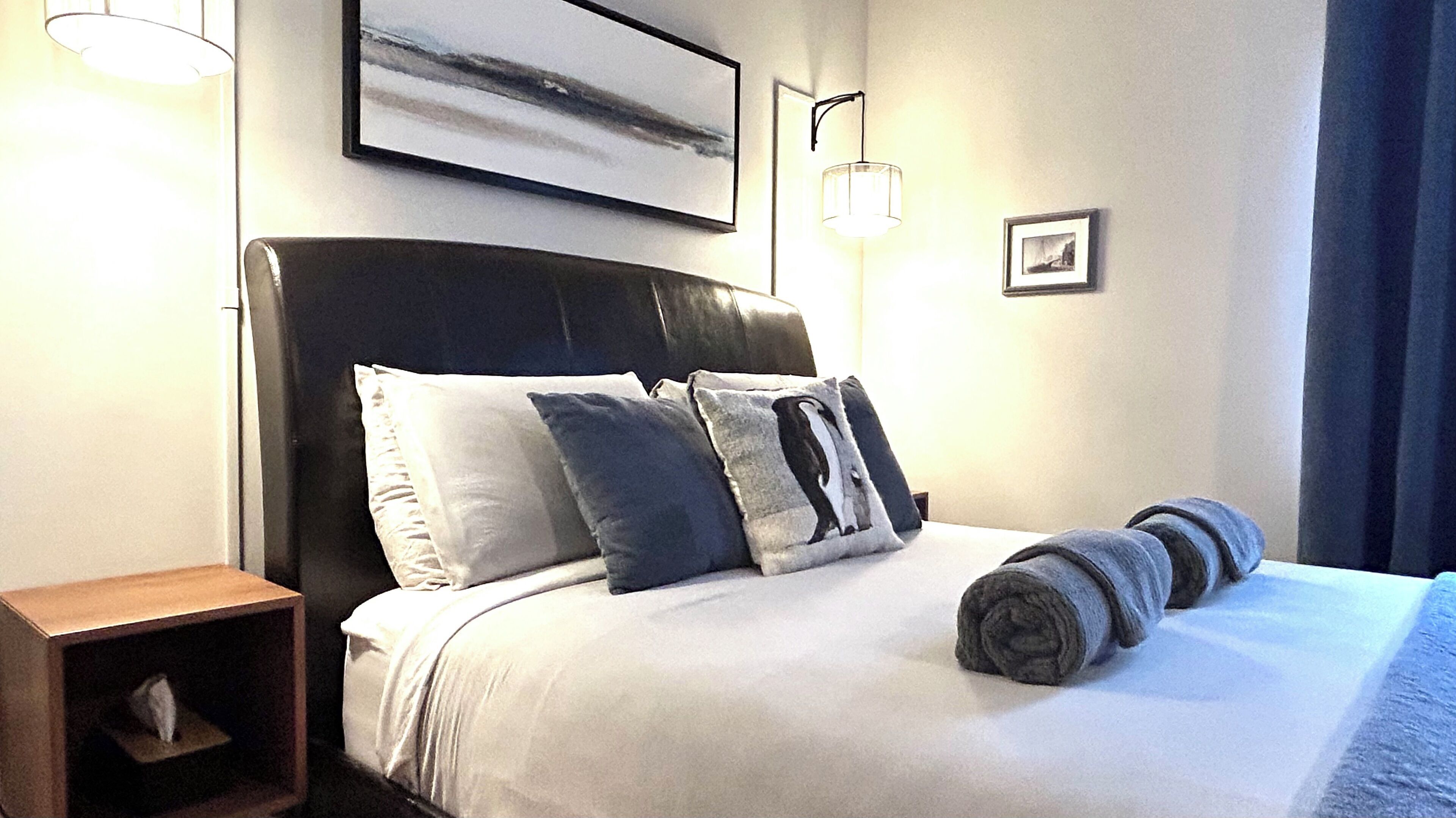 A neatly made bed with pillows and a rolled towel, a bedside table, and a wall-mounted picture.