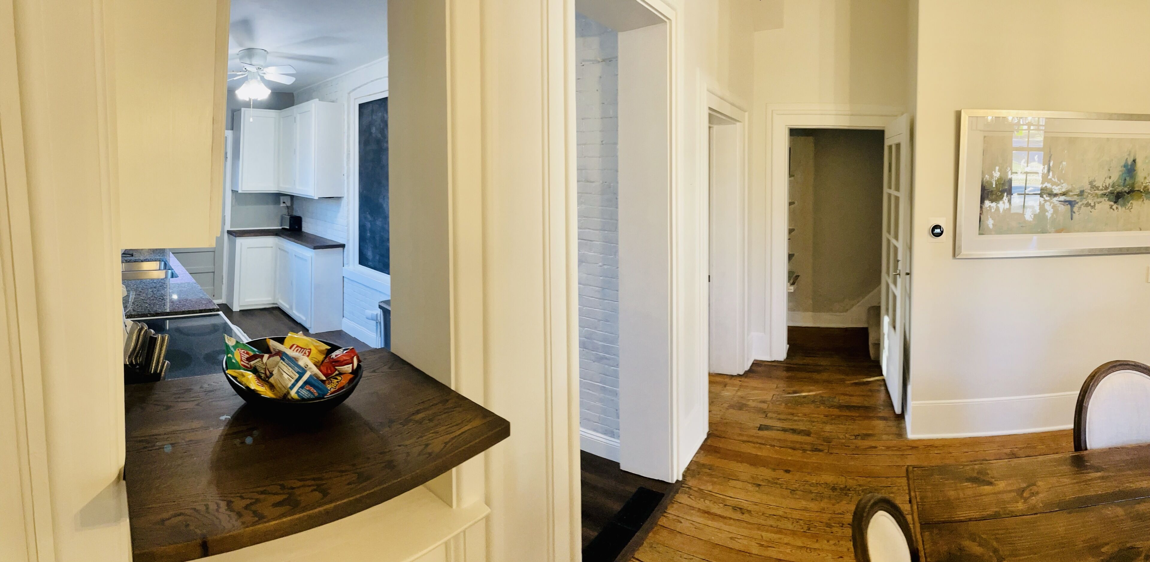 A hallway with a wooden floor, a dark brown countertop with a bowl of snacks, and a white door leading to a kitchen with white cabinets and a window.