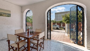 Un comedor con una mesa y sillas de madera, un cuadro en la pared y vistas a través de un portón abierto hacia una calle con coches y vegetación.