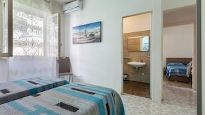 Un dormitorio con dos camas, una ventana adornada con cortinas decorativas, un cuadro en la pared y una puerta abierta que da a un baño.