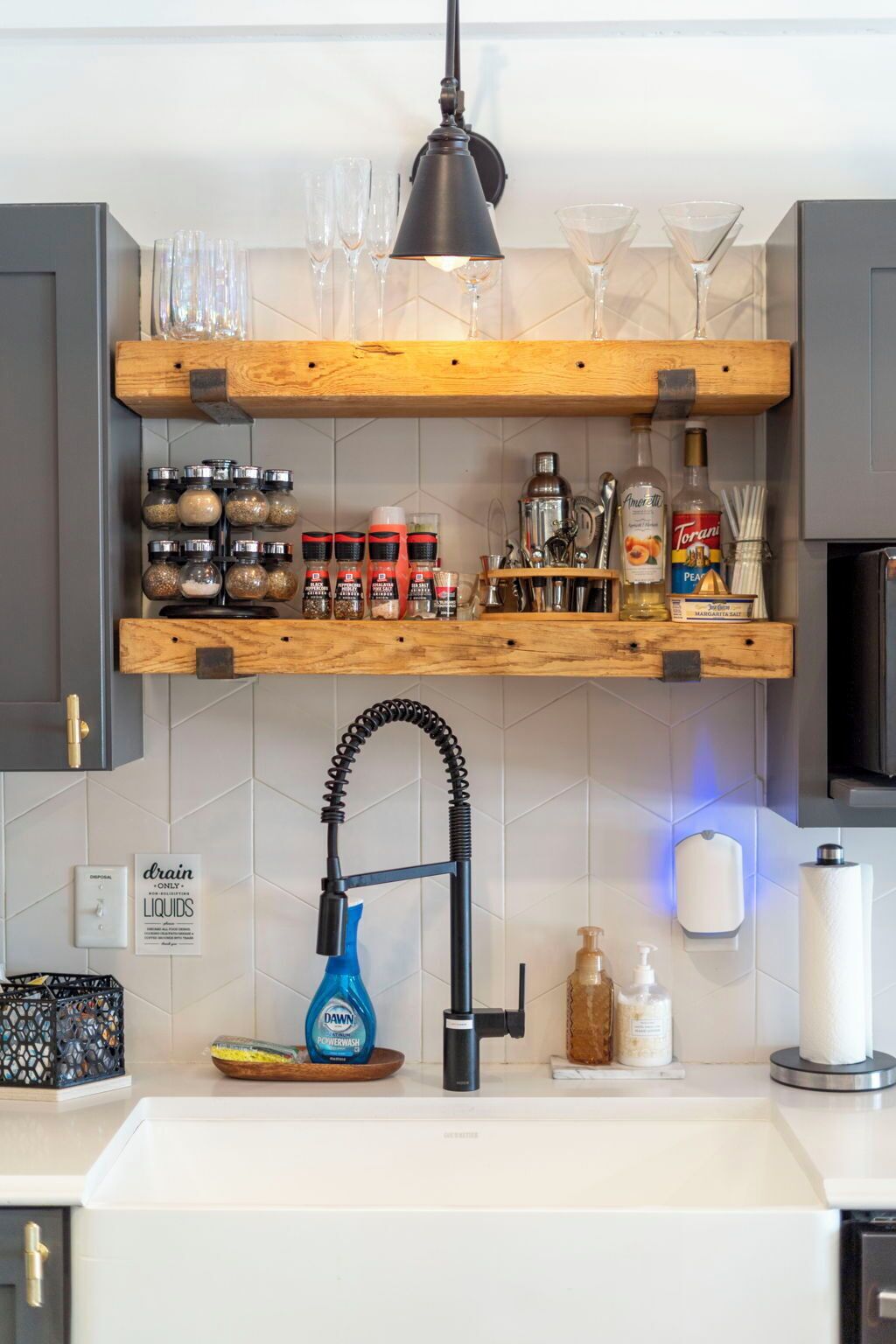 Eine Küche mit Holzböden, auf denen Gläser, Gewürze und Soßen stehen. Ein Spülbecken mit Wasserhahn und einer blauen Flasche Dawn Geschirrspülmittel ist sichtbar.
