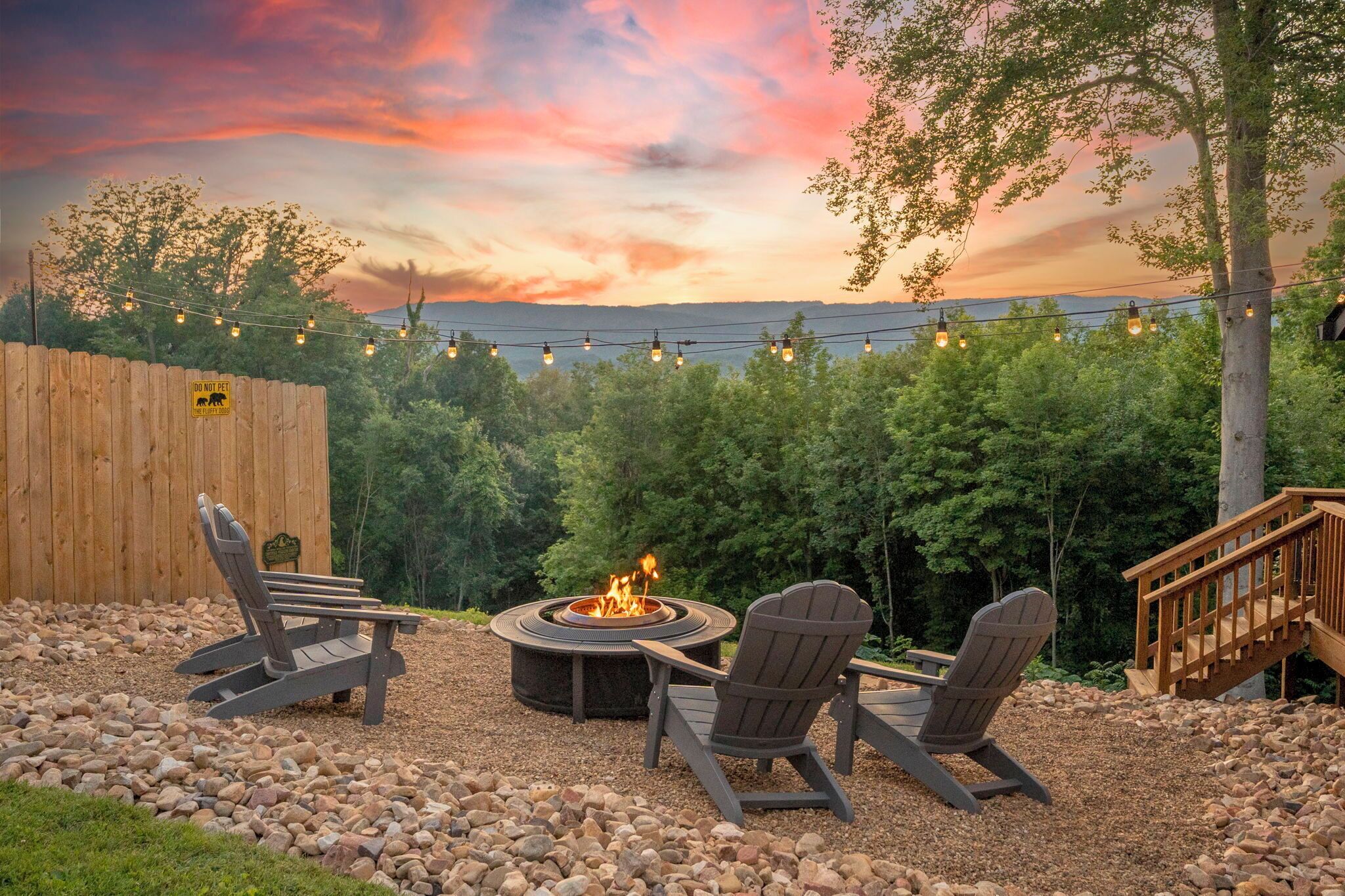 Patio con un hoyo para fuego, dos sillas Adirondack, una terraza de madera y guirnaldas de luces al atardecer.