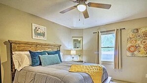 A bedroom with a bed, a ceiling fan, a window with curtains, and a wall with a framed picture.