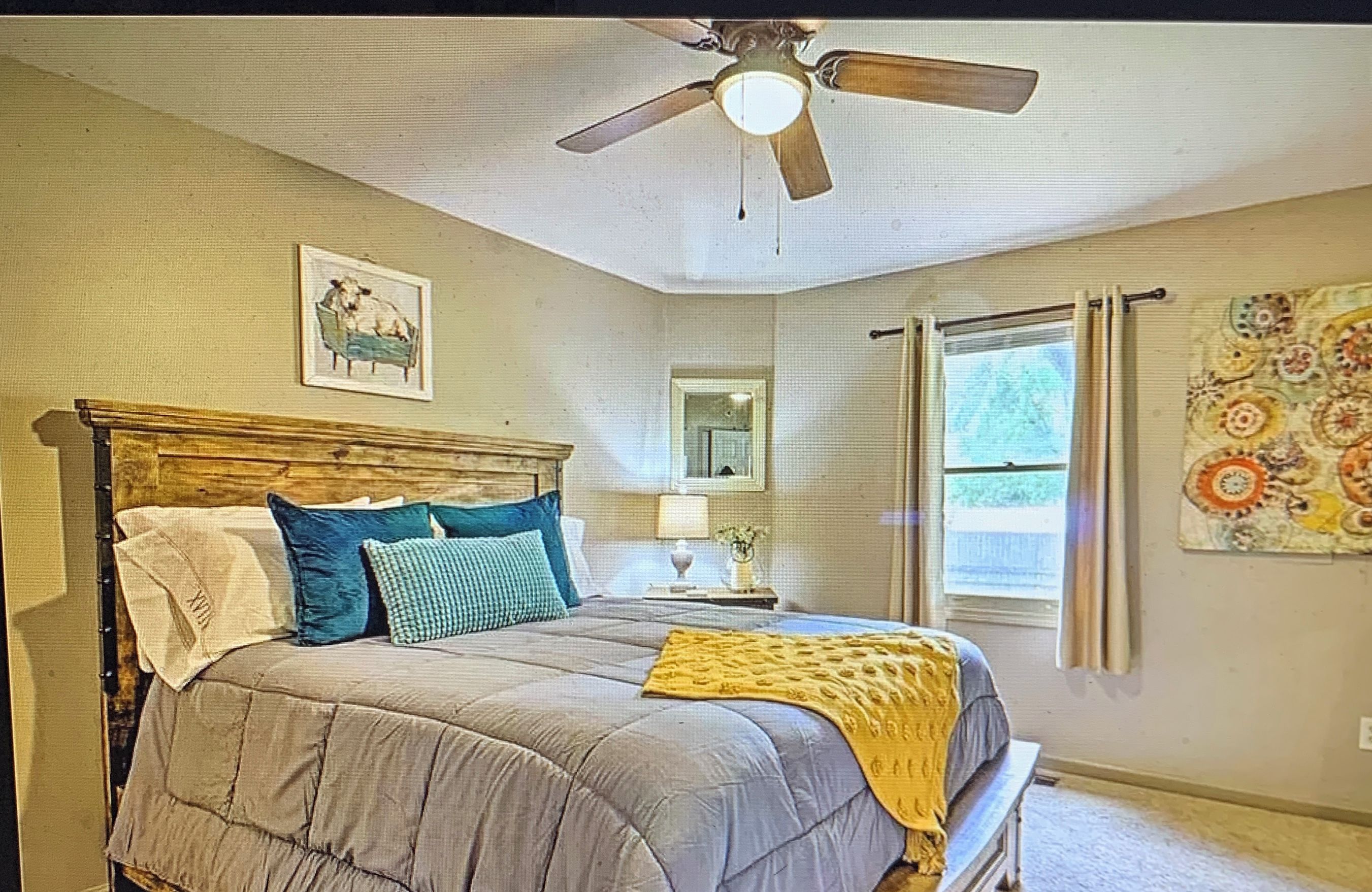Une chambre à coucher avec un lit, un ventilateur de plafond, une fenêtre avec des rideaux et un mur orné d’un tableau encadré.