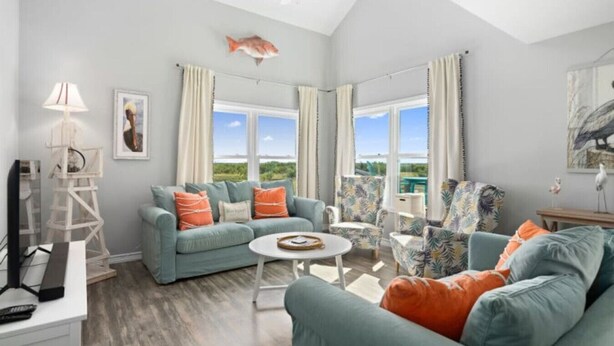 A spacious living room with a high ceiling, a large window offering a view of the outdoors, a ceiling fan, and a fish decoration. - Bayfront 2/2 Condo w/pool & Pier! Weekly Discounts (Rockport)