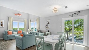 A spacious living room with a large sofa, dining table, and chairs. There is a view of the outdoors through large windows. - Bayfront 2/2 Condo w/pool & Pier! Weekly Discounts (Rockport)