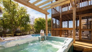 Una terraza de madera con un jacuzzi y piscina, rodeada de árboles y una zona vallada.