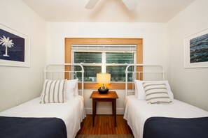 Una habitación con dos camas individuales, una mesa de madera con lámpara, una ventana con persianas y cuadros enmarcados en las paredes.