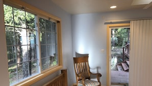 Quarto de canto com uma cadeira de balanço de madeira, janela com vista para árvores e porta que dá para o exterior.