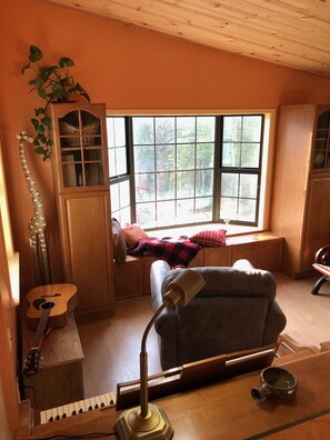 Una habitación acogedora con techo de madera, una ventana con vista a árboles, una estantería, una guitarra, un escritorio con lámpara y un sofá.