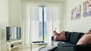 Een moderne woonkamer met een flatscreen televisie, een klein rond tafeltje, een bank en een groot raam met dunne gordijnen.