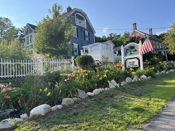 A cottage-style building with a white picket fence, a signboard with an American flag, and a garden with various flowers and rocks.