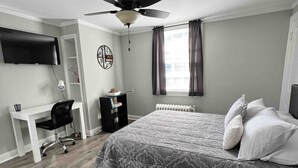 Un dormitorio con cama, escritorio, silla, ventilador de techo, televisor y radiador.