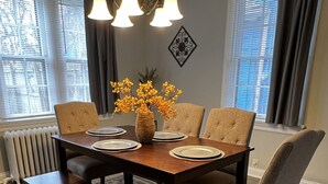 Un comedor con mesa de madera, cuatro sillas tapizadas, un banco, un florero con flores amarillas y una araña.