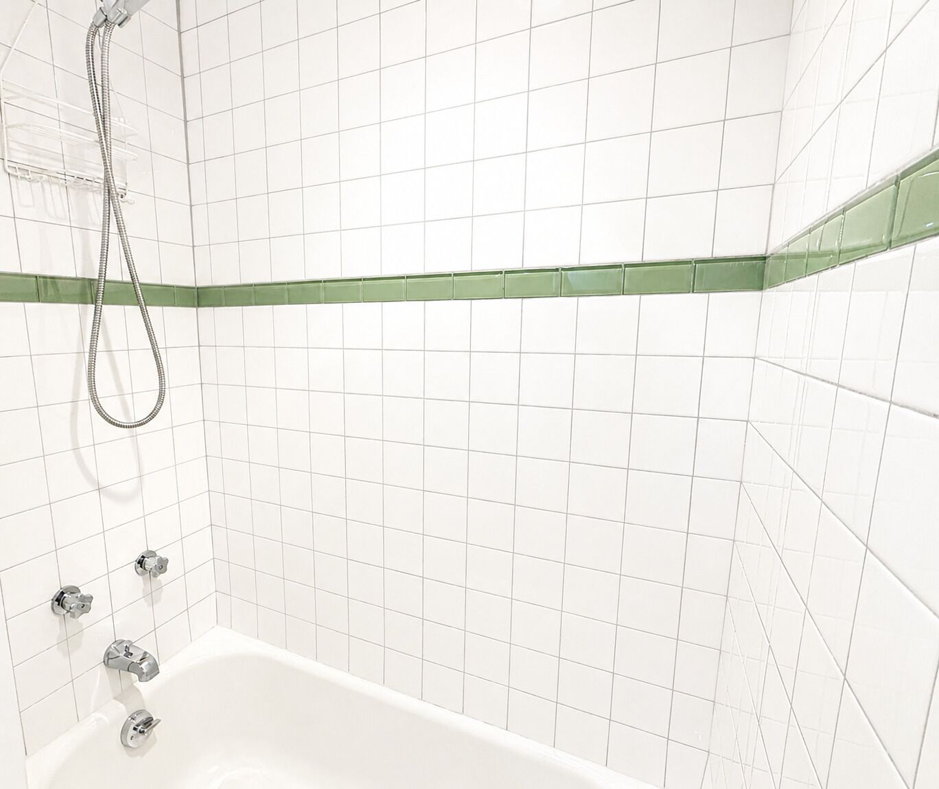 Une salle de bain avec des murs recouverts de carreaux blancs, une bande décorative verte et une baignoire dotée de robinetterie chromée.
